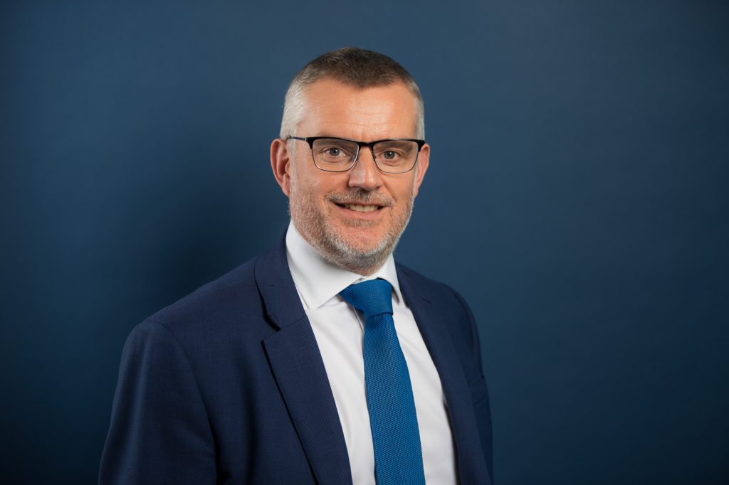 A man in a navy suit, white shirt, and blue tie smiles at the camera against a solid dark blue background. He has short hair, glasses, and a trimmed beard. New law firm headshots Manchester