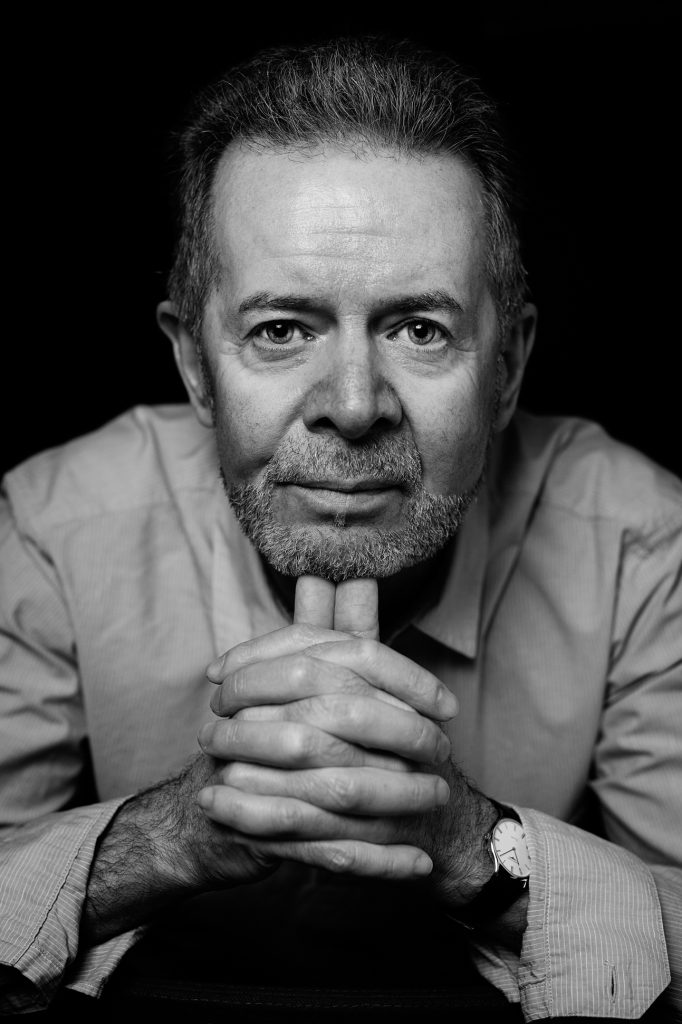 Black and white portrait of an older man with gray hair and beard, wearing a collared shirt and watch, resting his chin on his clasped hands, looking directly into the camera with a thoughtful expression. Writer at Manchester event
