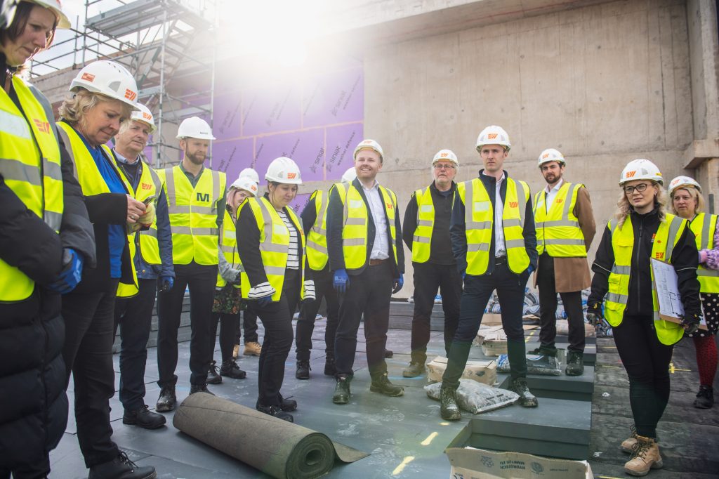 A group of people wearing yellow safety vests and white hard hats stand together at a construction site with unfinished concrete walls and scaffolding in the background. Sunlight shines from behind them. New construction site photography Manchester