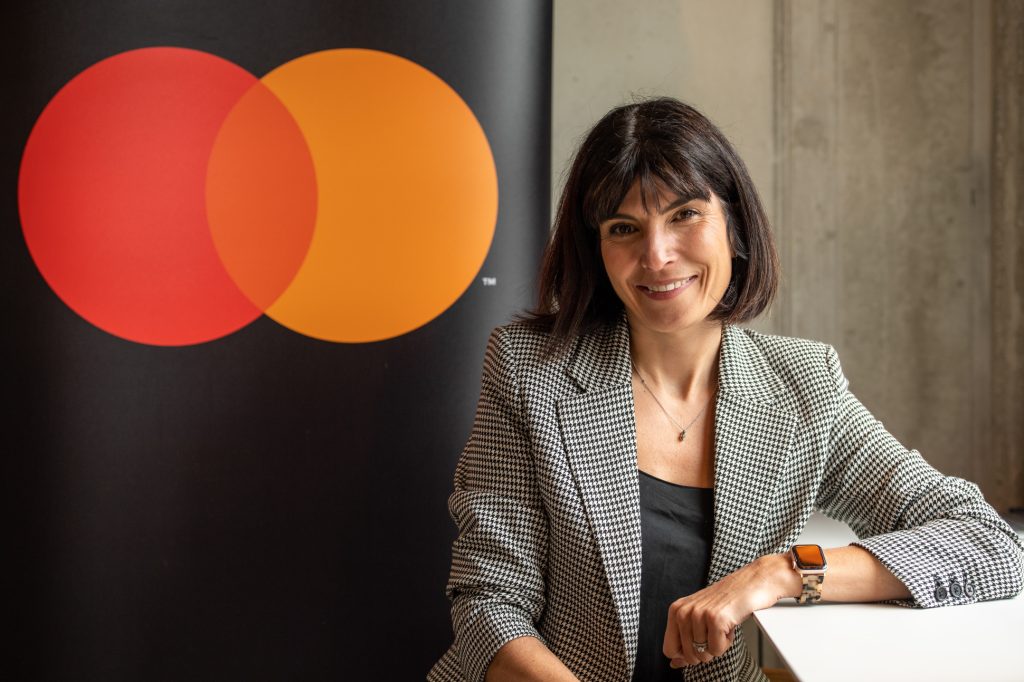 A woman in a checkered blazer sits at a white table, smiling, with a large Mastercard logo featuring overlapping red and orange circles visible in the background. corporate event photography Liverpool